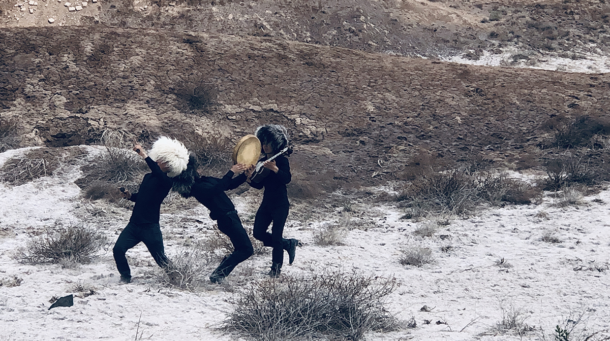 Three performers dressed in black, wearing shaggy black and white wigs, play instruments outside on scrubland.